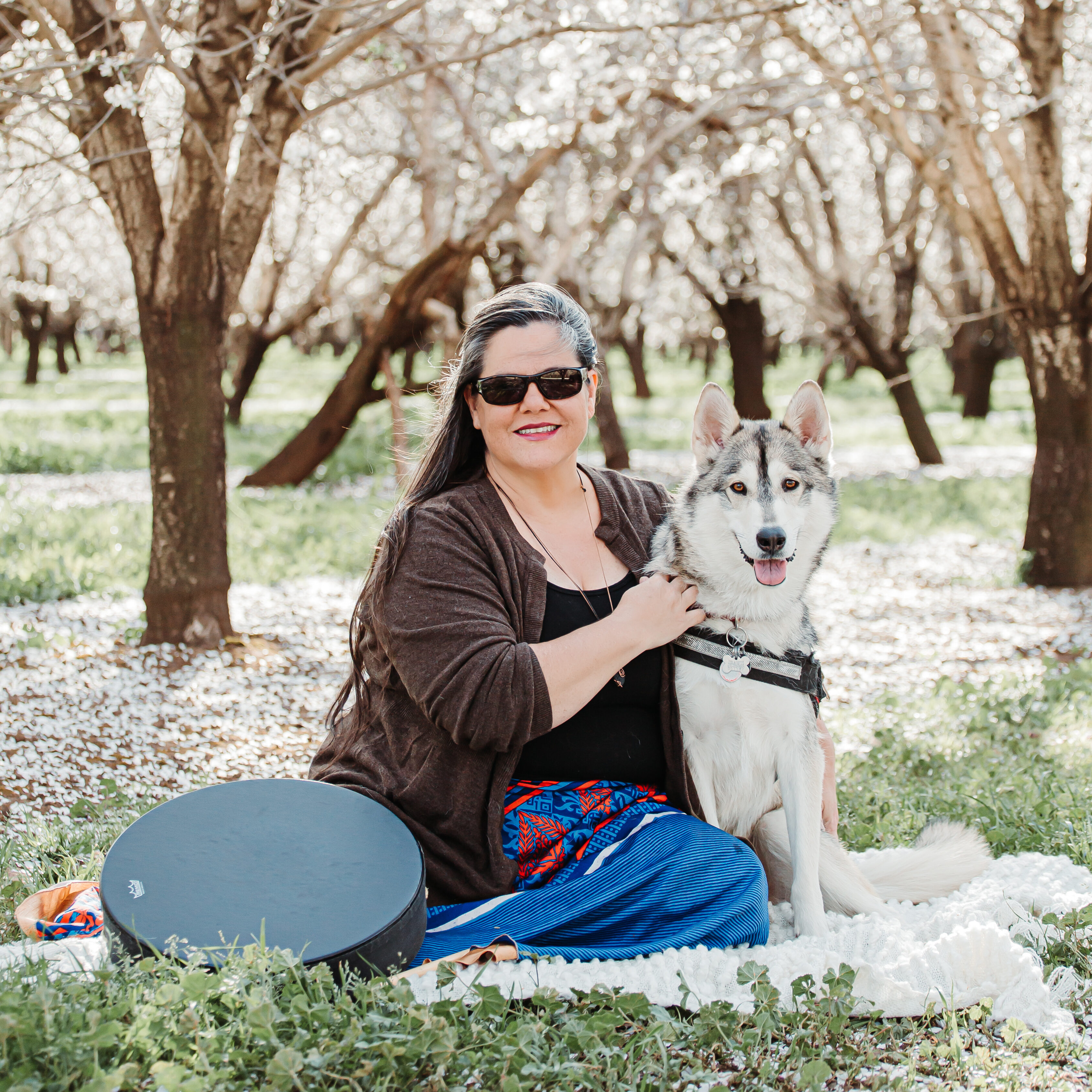 Amy and Rila sitting together in a blooming almond orchard with drum nearby.
