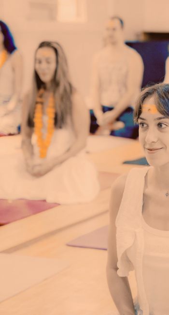 Woman smiling during a spiritual healing class, with yellow flower on forehead, sitting on yoga mat in a group setting.