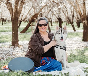 Amy and Rila sitting together in a blooming almond orchard with drum nearby.